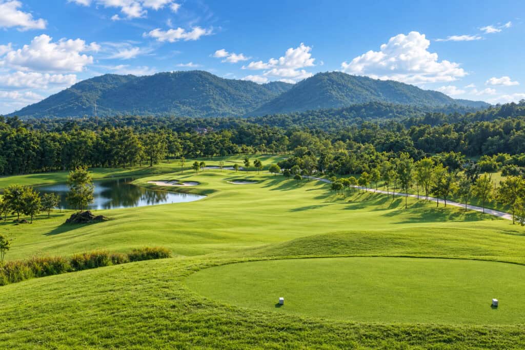 Chiangmai Highlands golf course with rolling fairways, bunkers and a lake beneath forested mountains and a blue sky.